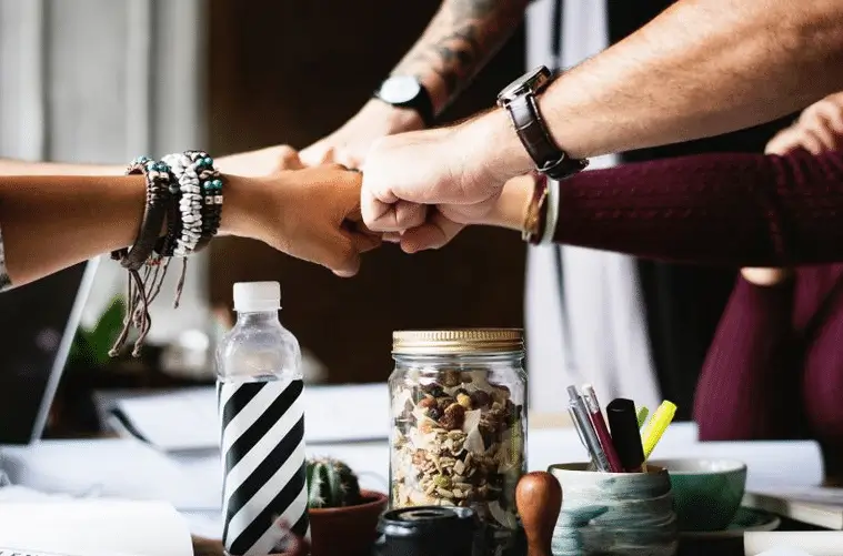 Fünf Hände treffen sich in einer Fist-Bump-Geste, umgeben von Büromaterial und Getränken auf einem Tisch.
