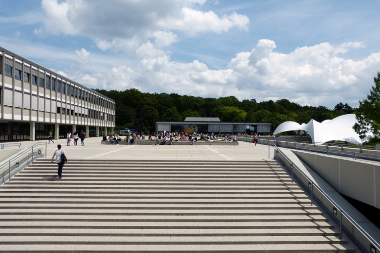A modern building with stairs, people in a square, and a green forest in the background under a partly cloudy sky.