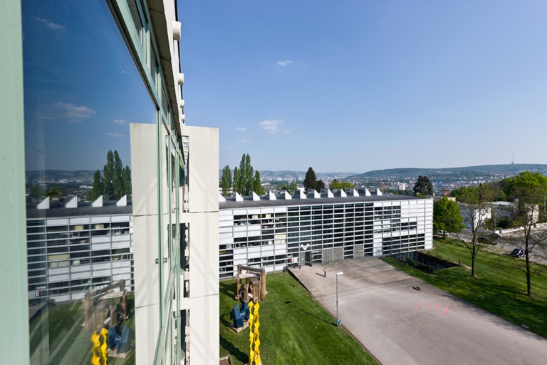 A modern building with large windows and an adjacent green space, set against an idyllic landscape under a clear sky.
