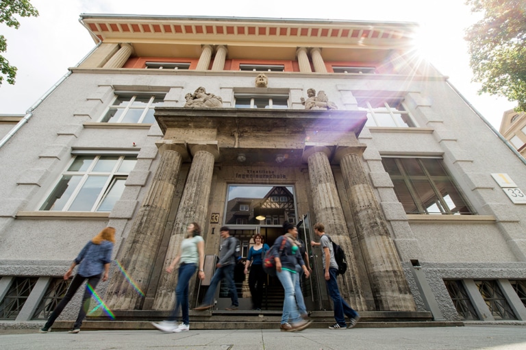 People enter the historic building, which features columns and sculptures on its facade.