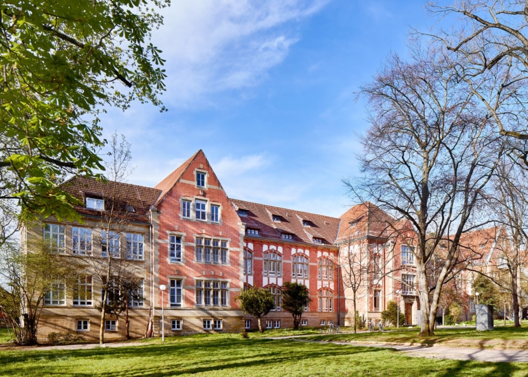 A historic building with a red facade and large windows, surrounded by trees and a green lawn.