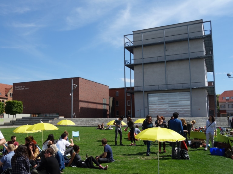 Menschen sitzen und stehen auf einer Wiese mit gelben Sonnenschirmen vor modernen Gebäuden an einem sonnigen Tag.