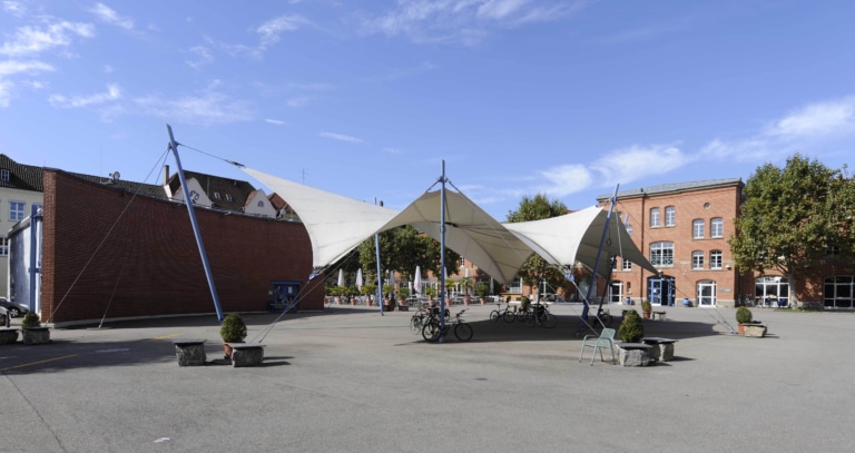 A clearing with a tent canopy and bicycles in front of historic buildings and trees under a blue sky.